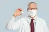 Elderly male doctor in glasses and mask holds a vaccine vial confidently. Wears a white coat and red tie, conveying professionalism and trust.