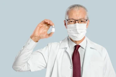Elderly male doctor in glasses and mask holds a vaccine vial confidently. Wears a white coat and red tie, conveying professionalism and trust.