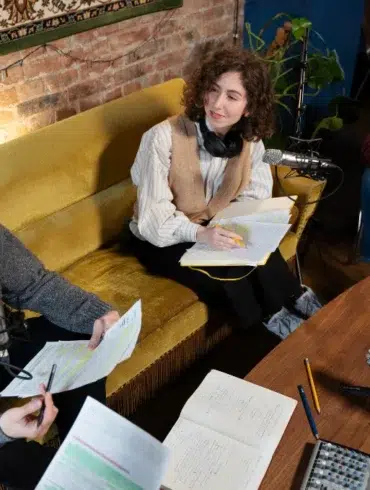 Three people are engaged in a discussion in a cozy room with a vintage rug and brick wall. They sit with papers and microphones, creating a podcast.