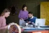 Three colleagues are working at a table with laptops and documents. One woman in pink writes in a notebook, while another in purple discusses with a man in blue, creating a focused and collaborative atmosphere.
