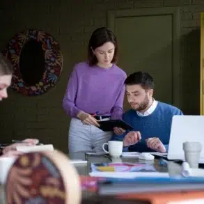 Three colleagues are working at a table with laptops and documents. One woman in pink writes in a notebook, while another in purple discusses with a man in blue, creating a focused and collaborative atmosphere.