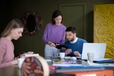 Three colleagues are working at a table with laptops and documents. One woman in pink writes in a notebook, while another in purple discusses with a man in blue, creating a focused and collaborative atmosphere.