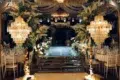 Elegant wedding venue with crystal chandeliers, floral decorations lining a glass runway, and banquet tables with white chairs and linens.