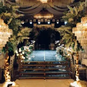 Elegant wedding venue with crystal chandeliers, floral decorations lining a glass runway, and banquet tables with white chairs and linens.