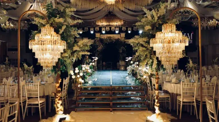 Elegant wedding venue with crystal chandeliers, floral decorations lining a glass runway, and banquet tables with white chairs and linens.