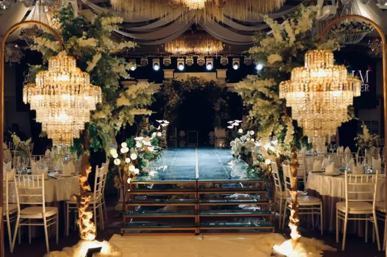 Elegant wedding venue with crystal chandeliers, floral decorations lining a glass runway, and banquet tables with white chairs and linens.