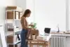 A woman in a brown sweater is packing items at a desk with a laptop, coffee cup, and boxes. Shelves with more boxes are in the background. Bright, organized space.