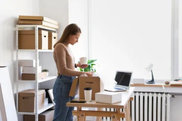 A woman in a brown sweater is packing items at a desk with a laptop, coffee cup, and boxes. Shelves with more boxes are in the background. Bright, organized space.