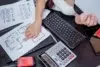 A person points to architectural blueprints on a desk, surrounded by a keyboard, calculator, red walkie-talkie, and tablet, creating a focused atmosphere.