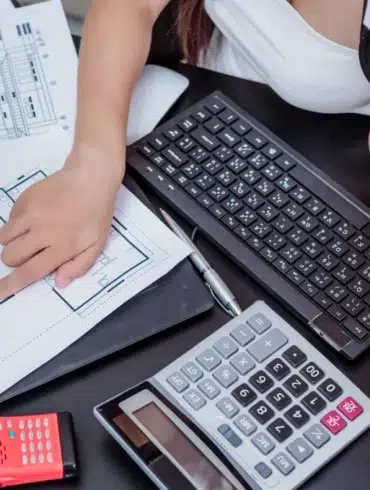 A person points to architectural blueprints on a desk, surrounded by a keyboard, calculator, red walkie-talkie, and tablet, creating a focused atmosphere.