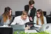 Four colleagues collaborate at a desk with laptops and documents, discussing work in a modern office setting.