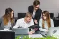 Four colleagues collaborate at a desk with laptops and documents, discussing work in a modern office setting.