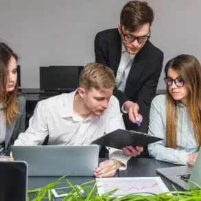 Four colleagues collaborate at a desk with laptops and documents, discussing work in a modern office setting.