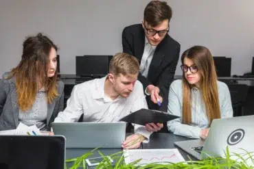 Four colleagues collaborate at a desk with laptops and documents, discussing work in a modern office setting.