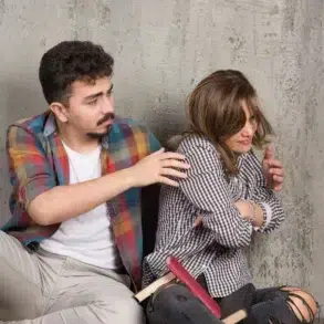 A young man reaching out to a distressed young woman sitting against a textured wall with books scattered around them.
