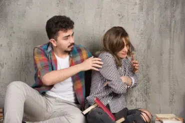 A young man reaching out to a distressed young woman sitting against a textured wall with books scattered around them.