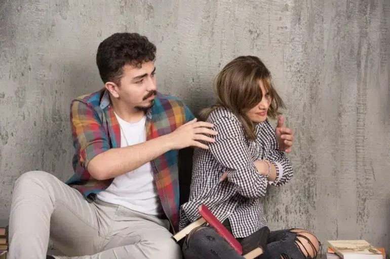 A young man reaching out to a distressed young woman sitting against a textured wall with books scattered around them.
