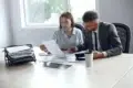 A man and a woman sit at a conference table, reviewing documents together. The office setting is bright and organized, conveying a focused teamwork atmosphere.