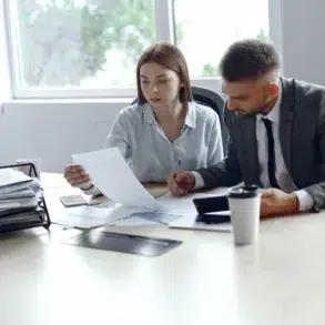 A man and a woman sit at a conference table, reviewing documents together. The office setting is bright and organized, conveying a focused teamwork atmosphere.
