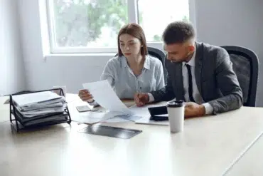 A man and a woman sit at a conference table, reviewing documents together. The office setting is bright and organized, conveying a focused teamwork atmosphere.