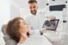 Dentist showing a dental X-ray on a tablet to a patient reclining in a dental chair in a bright clinic.