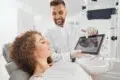 Dentist showing a dental X-ray on a tablet to a patient reclining in a dental chair in a bright clinic.