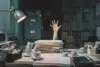 A cluttered desk piled high with papers. A hand reaching up from behind the stack suggests being overwhelmed, surrounded by cups and office supplies.