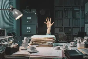 A cluttered desk piled high with papers. A hand reaching up from behind the stack suggests being overwhelmed, surrounded by cups and office supplies.