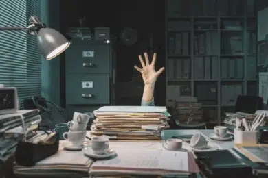 A cluttered desk piled high with papers. A hand reaching up from behind the stack suggests being overwhelmed, surrounded by cups and office supplies.