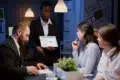A woman in a suit presents data on a tablet to engaged colleagues in an office. The room is softly lit, creating a focused and professional atmosphere.