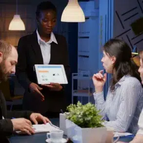 A woman in a suit presents data on a tablet to engaged colleagues in an office. The room is softly lit, creating a focused and professional atmosphere.