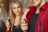 Young woman with long blonde hair smiling while holding a vape device, standing beside a man in a red plaid shirt, creating a relaxed and modern vibe.