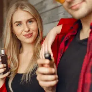 Young woman with long blonde hair smiling while holding a vape device, standing beside a man in a red plaid shirt, creating a relaxed and modern vibe.