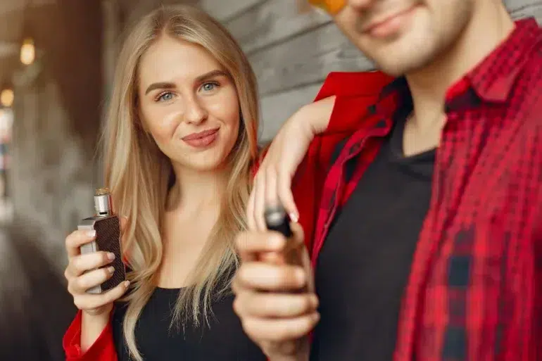 Young woman with long blonde hair smiling while holding a vape device, standing beside a man in a red plaid shirt, creating a relaxed and modern vibe.