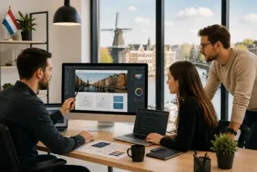 Three colleagues at a web design company in the Netherlands gather around a large desktop monitor and laptops to discuss a website interface design.