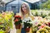 Person in a greenhouse holding two potted flowering plants, one with red flowers and the other with white flowers.