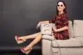 A woman in a colorful zigzag dress and sunglasses lounges on a beige sofa. She wears red wedge sandals and holds a "WOW!" graphic handbag, conveying a trendy, relaxed vibe.