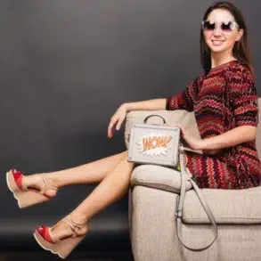 A woman in a colorful zigzag dress and sunglasses lounges on a beige sofa. She wears red wedge sandals and holds a "WOW!" graphic handbag, conveying a trendy, relaxed vibe.