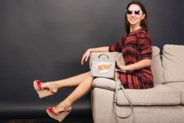 A woman in a colorful zigzag dress and sunglasses lounges on a beige sofa. She wears red wedge sandals and holds a "WOW!" graphic handbag, conveying a trendy, relaxed vibe.