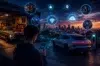 A person holds a smartphone near a sleek electric car parked outside a garage. Digital icons illustrate connectivity. City skyline at sunset in the background.
