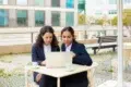 Two women in business attire focus intently on a laptop at an outdoor café table. Coffee cups are in front of them, with modern buildings in the background.