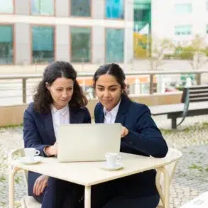 Two women in business attire focus intently on a laptop at an outdoor café table. Coffee cups are in front of them, with modern buildings in the background.