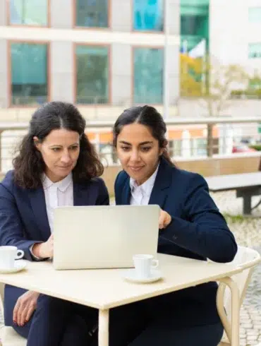 Two women in business attire focus intently on a laptop at an outdoor café table. Coffee cups are in front of them, with modern buildings in the background.