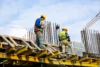 Construction workers in safety gear work on a high platform, surrounded by steel rods, against a cloudy sky, conveying an industrious tone.