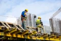 Construction workers in safety gear work on a high platform, surrounded by steel rods, against a cloudy sky, conveying an industrious tone.