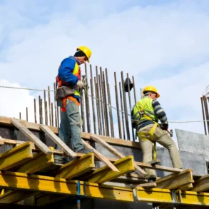 Construction workers in safety gear work on a high platform, surrounded by steel rods, against a cloudy sky, conveying an industrious tone.