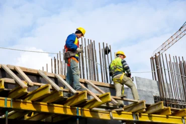 Construction workers in safety gear work on a high platform, surrounded by steel rods, against a cloudy sky, conveying an industrious tone.