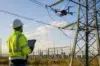An engineer wearing a white hard hat and a high-visibility safety jacket uses a tablet to monitor a drone power line inspection near a large electrical pylon.