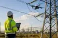 An engineer wearing a white hard hat and a high-visibility safety jacket uses a tablet to monitor a drone power line inspection near a large electrical pylon.