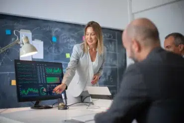 A woman in a gray suit gestures at a computer screen displaying financial graphs during a business meeting. Colleagues listen intently, with a chalkboard in the background.